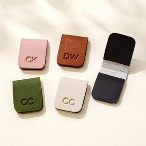 Puede incluir: Cinco marcadores magn&eacute;ticos de cuero en varios colores: rosa, marr&oacute;n, verde, blanco roto y negro. Cada marcador presenta iniciales en relieve: CK, DW, CC y CC. Los marcadores son rectangulares con un dise&ntilde;o plegado.