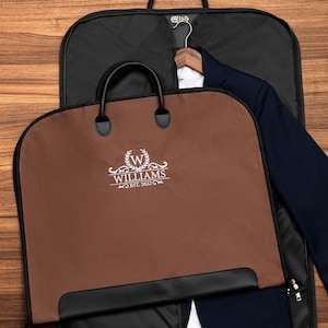 Peut inclure: Un sac à vêtements marron avec des bordures et des poignées noires, avec le nom "WILLIAMS" et "EST. 2022" en écriture blanche. Une veste de costume bleu marine est suspendue à l'intérieur d'un sac à vêtements noir, sur une surface en bois.