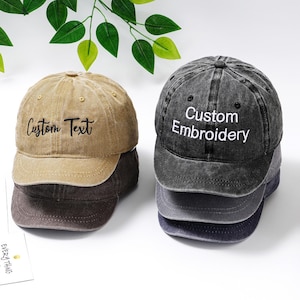 Könnte beinhalten: Eine Reihe von Baseballkappen in Beige-, Grau- und Marinefarben. Die beige Kappe ist mit "Custom Text" bestickt, während die graue Kappe "Custom Embroidery" zeigt. Die Kappen haben einen verwaschenen, Vintage-Look.