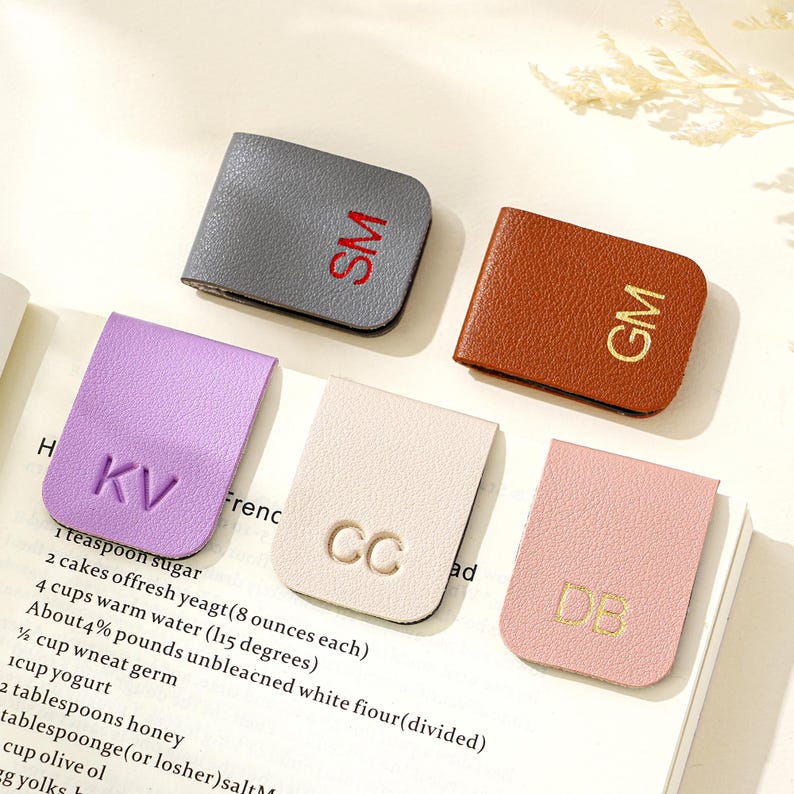 Puede incluir: Cinco marcadores magn&eacute;ticos de cuero en varios colores: gris con "SM" rojo, marr&oacute;n con "GM" dorado, lavanda con "KV", crema con "CC" y rosa con "DB". Los marcadores est&aacute;n sobre una p&aacute;gina de libro.