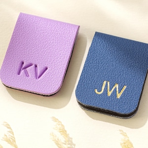 Puede incluir: Dos marcap&aacute;ginas de cuero, uno lila y otro azul. El marcap&aacute;ginas lila tiene las iniciales "KV" en relieve, mientras que el marcap&aacute;ginas azul presenta las iniciales "JW" bordadas. Ambos son rectangulares con bordes redondeados.