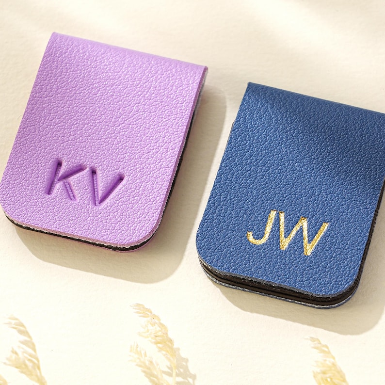 Puede incluir: Dos marcap&aacute;ginas de cuero, uno lila y otro azul. El marcap&aacute;ginas lila tiene las iniciales "KV" en relieve, mientras que el marcap&aacute;ginas azul presenta las iniciales "JW" bordadas. Ambos son rectangulares con bordes redondeados.