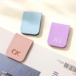 Puede incluir: Tres marcap&aacute;ginas personalizados de cuero en colores pastel: azul claro, rosa y lavanda. Cada marcap&aacute;ginas presenta iniciales bordadas: "TC", "CK" y "AS". Los marcap&aacute;ginas tienen un borde negro.