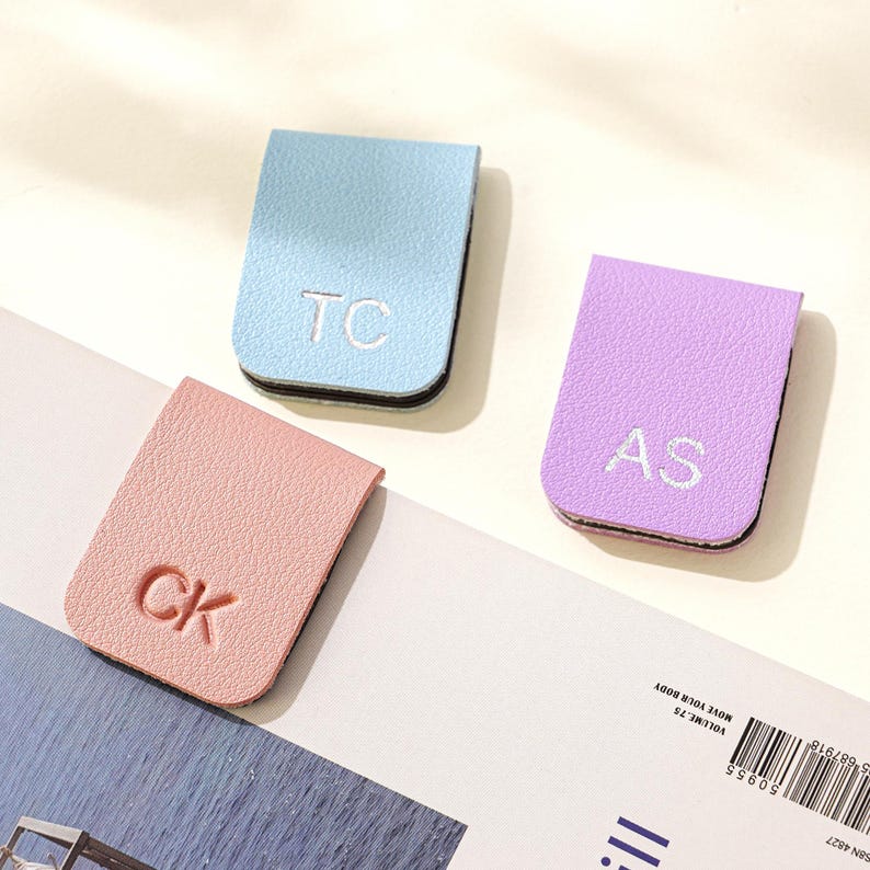 Puede incluir: Tres marcap&aacute;ginas personalizados de cuero en colores pastel: azul claro, rosa y lavanda. Cada marcap&aacute;ginas presenta iniciales bordadas: "TC", "CK" y "AS". Los marcap&aacute;ginas tienen un borde negro.