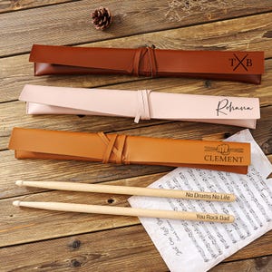 May include: Three leather drumstick cases in brown, pink, and tan, each with a tie closure. The cases are personalized with initials, a name, and a name with a drum graphic. Two wooden drumsticks with the text "No Drums No Life" and "You Rock Dad" rest on sheet music.