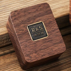 May include: A wooden box with a dark brown finish. A rectangular metal plaque with the initials "R&S" and the date "08.23.26" is affixed to the top. The box is set against a wooden surface.