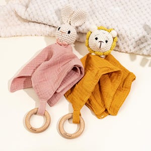 May include: Two baby comforters, one pink and one mustard yellow, each with a crocheted animal head and a wooden ring. The pink comforter has a bunny head, and the yellow one has a lion head. The wooden rings are engraved with names.