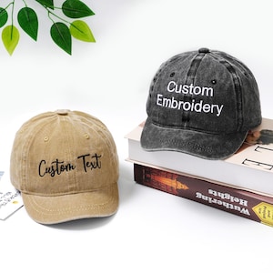 Pode incluir: Duas bonés de beisebol, uma bege e outra cinza escuro, com texto bordado. O boné bege diz "Custom Text" e o cinza "Custom Embroidery". Ambos os bonés estão sobre uma superfície branca.