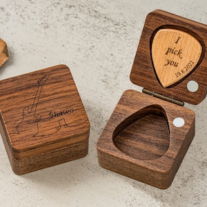 May include: A wooden box with a guitar design on the lid and the name "Shawn" engraved. The box is open and reveals a small wooden pick with the text "I pick you 19.6.2021" engraved on it.