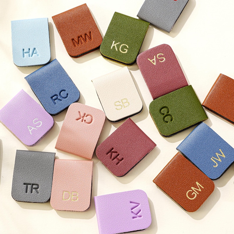 Puede incluir: Una colecci&oacute;n de clips para billetes de cuero en varios colores, como azul, verde y rosa. Cada clip presenta iniciales en relieve en dorado o del mismo color. Los clips rectangulares tienen un borde superior curvado.