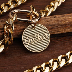 May include: A gold-colored dog collar with a thick chain design. The collar features a round tag with the name "Tucker" in a cursive font. The tag has a striped background. The collar is displayed on a wooden surface.