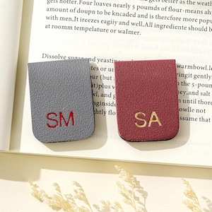 Puede incluir: Dos marcap&aacute;ginas de cuero. Uno es gris con las iniciales bordadas en rojo "SM", el otro es burdeos con las iniciales bordadas en oro "SA". Los marcadores son rectangulares con un borde superior curvo.