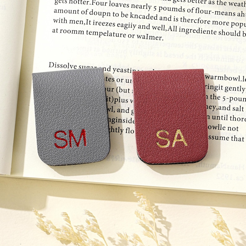 Puede incluir: Dos marcap&aacute;ginas de cuero. Uno es gris con las iniciales bordadas en rojo "SM", el otro es burdeos con las iniciales bordadas en oro "SA". Los marcadores son rectangulares con un borde superior curvo.