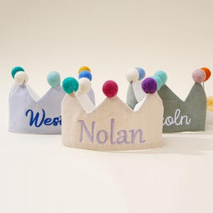 Könnte beinhalten: Stoffkronen in Pastelltönen, jede mit einem Namen in einer kontrastierenden Farbe bestickt. Die Kronen sind mit bunten Pompons verziert. Die mittlere Krone ist beige mit dem Namen "Nolan" in Lila gestickt.