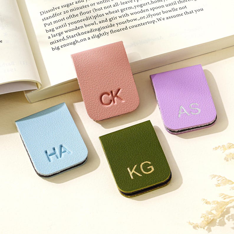 Puede incluir: Cuatro sujetap&aacute;ginas de cuero en colores pastel: azul claro, rosa, verde y morado. Cada uno tiene iniciales diferentes: HA, CK, KG y AS. Los accesorios para libros est&aacute;n sobre una superficie blanca junto a un libro abierto.