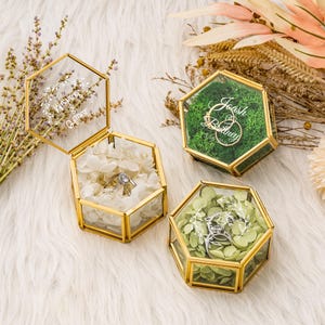 Könnte beinhalten: Drei sechseckige Glas-Ringboxen mit goldfarbenem Rand. Eine Box ist geöffnet und zeigt einen Diamantring auf weißen Blütenblättern. Die anderen beiden Boxen enthalten Moos und Grün. Die offene Box trägt den Text "Whitney & Owen".