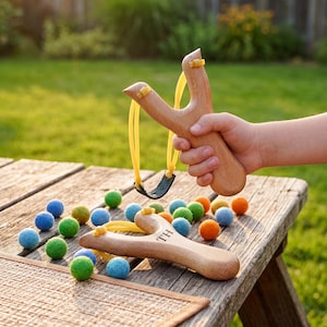 Könnte beinhalten: Zwei Holzschleudern mit gelben Gummibändern und schwarzen Ledertaschen. Eine Schleuder wird in der Hand gehalten, während die andere auf einer Holzoberfläche neben zahlreichen kleinen, bunten Filzkugeln in Grün, Blau und Orange liegt.