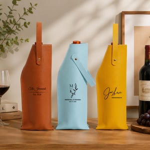 Pode incluir: Três porta-garrafas de vinho verticais nas cores castanho, azul claro e amarelo, cada um com uma alça e texto preto personalizado. O porta-garrafas castanho diz The Jeremiah Est. 2026, o azul mostra MK com um desenho de folha e o amarelo diz Joshua.