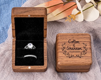 Peut inclure: Une boîte à bague en bois avec une finition brun foncé. Le couvercle est gravé des noms "Colton & Shannon" et de la date "06.03.2023". La boîte est ouverte et montre un intérieur en velours avec une bague et une alliance.