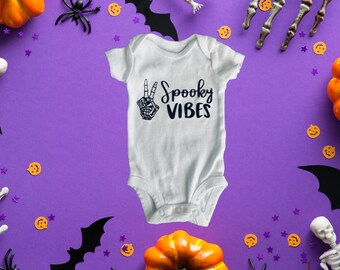 May include: A white baby grow with a black skeleton hand making a peace sign and the text "Spooky Vibes". The grow is surrounded by Halloween decorations, including pumpkins, bats, spiders, and confetti.