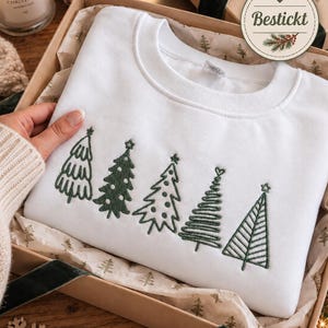Könnte beinhalten: Ein weißes Sweatshirt mit Rundhalsausschnitt und fünf verschiedenen, dunkelgrün gestickten Weihnachtsbäumen auf der Brust. Jeder Baum hat ein einzigartiges Muster aus Linien, Punkten und Sternen. Das Sweatshirt liegt in einer Geschenkbox.