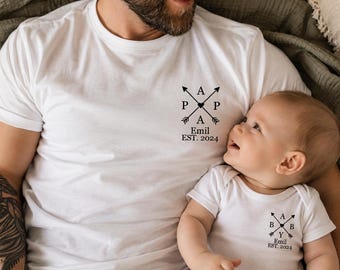 May include: A white short-sleeved t-shirt and a white short-sleeved baby bodysuit. The t-shirt features a black graphic of crossed arrows with the letters P, A, P, A and the text Emil EST. 2024. The bodysuit has crossed arrows with B, A, B, Y and Emil EST. 2024.