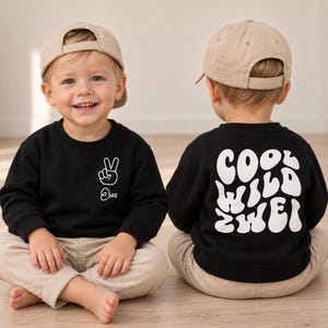 Może przedstawiać: Czarna bluza z długim rękawem z białym znakiem pokoju i imieniem Elias na lewej piersi oraz napisem COOL WILD ZWEI dużymi, białymi, falistymi literami na plecach, zestawiona z beżowymi spodniami i beżową czapką z daszkiem.