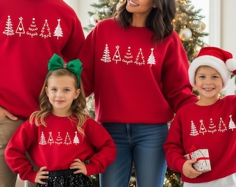 May include: A family of four wearing matching red sweatshirts with a white Christmas tree design. The family is standing in front of a Christmas tree and fireplace, with gifts and stockings visible. The girl is wearing a black sequin skirt and green bow.