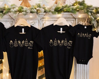 May include: Four black Christmas t-shirts and a baby bodysuit hang on wooden hangers. Each garment has a gold outline of festive characters. The shirts are labelled "Mama", "Papa", "Baby", and "Mia".