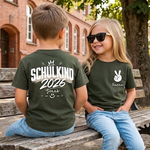 May include: Two olive green t-shirts. The back of one shirt features a crown icon, the text "SCHULKIND 2026", the name "Jonas", and a smiley face. The front of the other shirt displays a white peace sign graphic and the name "Hanna".