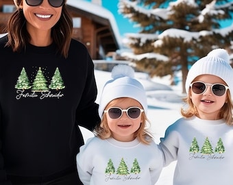 May include: Black and white sweatshirts with a watercolor Christmas tree design. The sweatshirts have the text "Familie Schneider" in a script font. The image shows a family wearing the sweatshirts in a snowy outdoor setting.