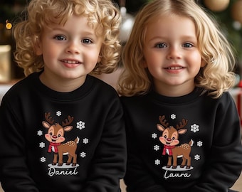 May include: Two children wearing matching black sweatshirts with a reindeer graphic and snowflakes. The reindeer has a red scarf. The name "Daniel" is printed on one sweatshirt, and "Laura" on the other. The background includes a Christmas tree and wrapped gifts.