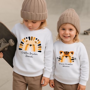 Puede incluir: Dos niños con sudaderas blancas y gorros beiges. Una sudadera presenta un diseño de león con el texto "Großer Bruder Daniel". La otra tiene un diseño de tigre con el texto "Kleine Schwester Johanna".