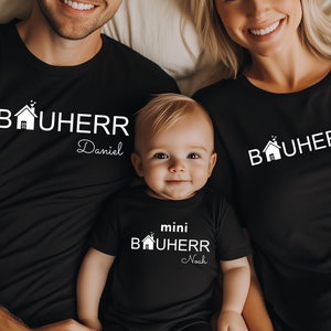 May include: A family of three wearing black t-shirts with a white house graphic and the text "Bauherr" with their names underneath. The father's shirt says "Daniel", the mother's shirt says "Johanna", and the baby's shirt says "mini Bauherr Noah".