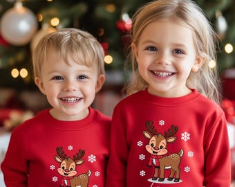May include: Two children wearing red sweatshirts with a reindeer graphic and snowflakes. The reindeer has a red scarf. The name "Daniel" is printed on one sweatshirt, and "Laura" on the other. A Christmas tree is in the background.