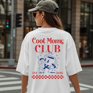 Pode incluir: Camiseta branca com o texto "Cool Moms Club" em vermelho e um desenho animado de uma xícara de café azul. A camisa também apresenta o texto "EST. 2026" e um padrão xadrez vermelho e branco.