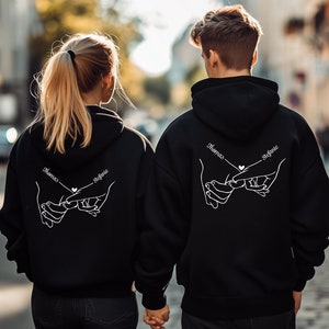 Könnte beinhalten: Zwei schwarze Hoodies mit weißen Umrissen von Händen, die sich die kleinen Finger halten. Der Text "Thomas" und "Sophie" steht über den Händen.
