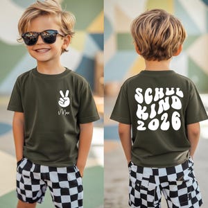 Könnte beinhalten: Ein olivgrünes T-Shirt mit einem weißen Peace-Zeichen und dem Namen Max auf der Vorderseite sowie dem Text SCHUL KIND 2026 in weißer, geschwungener Schrift auf der Rückseite, kombiniert mit schwarz-weiß karierten Shorts und einer schwarzen Sonnenbrille.