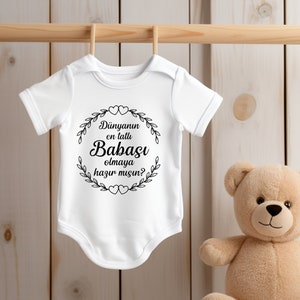 May include: White baby onesie with black text that reads "Dunyannın en tatlı Babası olmaya hazır mısın?"  The onesie is hanging on a wooden hanger. A brown teddy bear is sitting on a white stool.