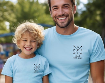 May include: Two people wearing matching light blue t-shirts with a black arrow design and the text "Emil Est. 2017". The person on the left is a child and the person on the right is an adult.