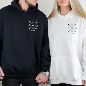 Könnte beinhalten: Passende Kapuzenpullover in Schwarz und Weiß. Jeder hat eine Vordertasche und ein weißes Grafikdesign aus gekreuzten Pfeilen, Initialen und einem Datum. Der schwarze Pullover ist links, der weiße rechts.