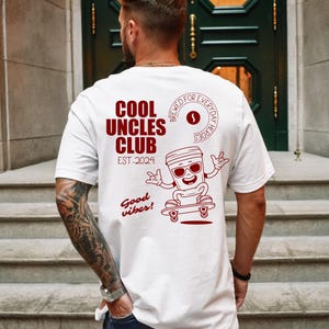 Könnte beinhalten: Weißes T-Shirt mit roter Grafik auf dem Rücken. Die Grafik zeigt den Text "COOL UNCLES CLUB EST. 2024" und eine Cartoon-Kaffeetasse auf einem Skateboard mit Sonnenbrille. Das Shirt hat auch die Worte "Brewed for everyday heroes" und "Good vibes!"
