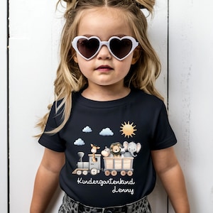 Könnte beinhalten: Ein marineblaues Kurzarm-T-Shirt mit einem Zugmotiv, das Zeichentricktiere transportiert, darunter eine Giraffe, ein Löwe und ein Elefant. Der Text "Kindergartenkind Lenny" befindet sich unter dem Zug, mit Sonne und Wolken im Hintergrund.