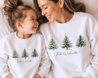 May include: White sweatshirts with matching Christmas tree designs. Each sweatshirt features three decorated trees and the text "Frohe Weihnachten!" in black script. The sweatshirts are worn by a mother and child.