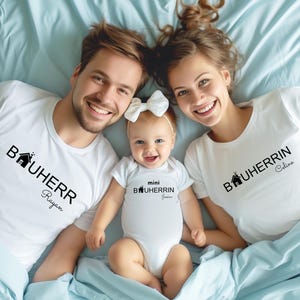 Könnte beinhalten: Eine dreiköpfige Familie trägt weiße T-Shirts mit dem Text "Bauherr" und einem Haussymbol. Das Shirt des Vaters lautet "Bauherr Rayan", das Shirt der Mutter lautet "Bauherr Celine" und das Shirt des Babys lautet "mini Bauherr Jessie".