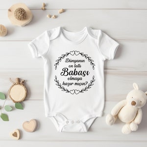 May include: White baby onesie with black text that reads "D&uuml;nyanın en tatlı Babası olmaya hazır mısın?"  The text is surrounded by a wreath of leaves and hearts.