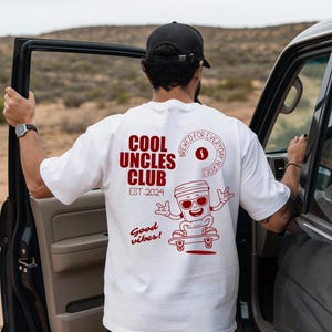 Könnte beinhalten: Weißes T-Shirt mit roter Grafik eines Kaffeepot auf einem Skateboard, mit dem Text "COOL UNCLES CLUB EST. 2024" und "Good vibes!". Das T-Shirt wird von einer Person neben einem Auto getragen.