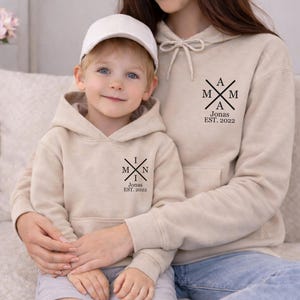 May include: Beige hoodies with black text that reads "I M N Jonas EST. 2022" and "A M M Jonas EST. 2022". The child is wearing a white baseball cap. The hoodies have a drawstring and a front pocket. The child is wearing grey shorts.