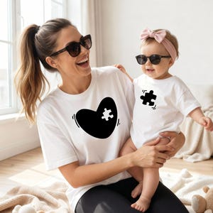 Könnte beinhalten: Ein weißes T-Shirt mit einem großen schwarzen Herz-Motiv und einem weißen Puzzleteil-Ausschnitt, kombiniert mit einem passenden weißen Baby-Body, der ein einzelnes schwarzes Puzzleteil zeigt. Beide Kleidungsstücke bestehen aus weißem Baumwollstoff.
