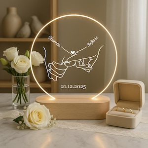 Può includere: Una decorazione nuziale circolare in acrilico illuminato con base in legno. Il design presenta due mani che si tengono, con i nomi "Thomas" e "Stefanie" e la data "21.12.2025". Una scatola per anelli con due fedi d'oro è accanto alla lampada.